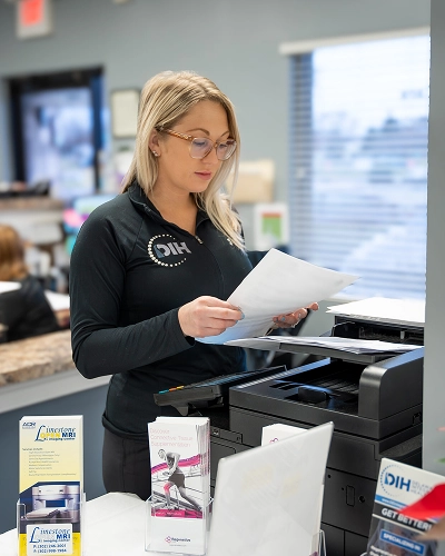 Woman wearing glasses and a black shirt with DIH logo printing on her chest standing in front of a printer holding a paper sheet.