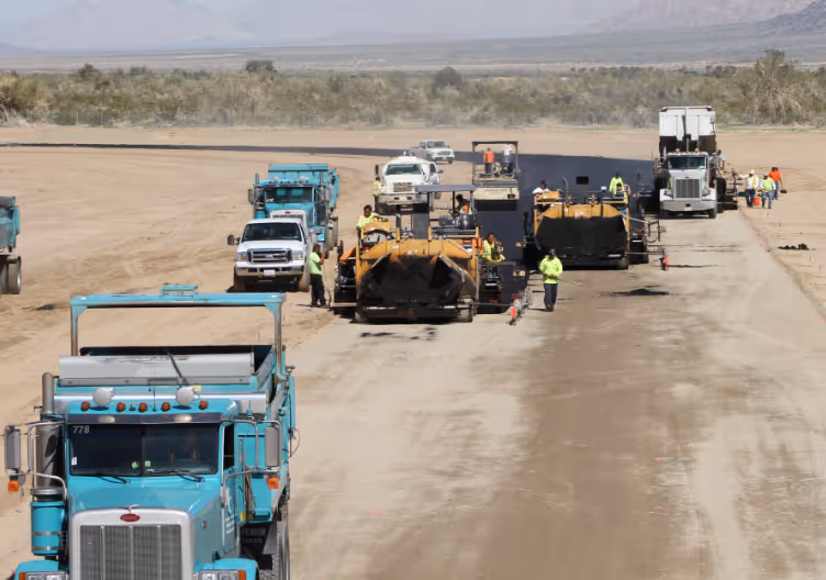 Construction workers and heavy machinery paving a long stretch of road in a desert landscape.