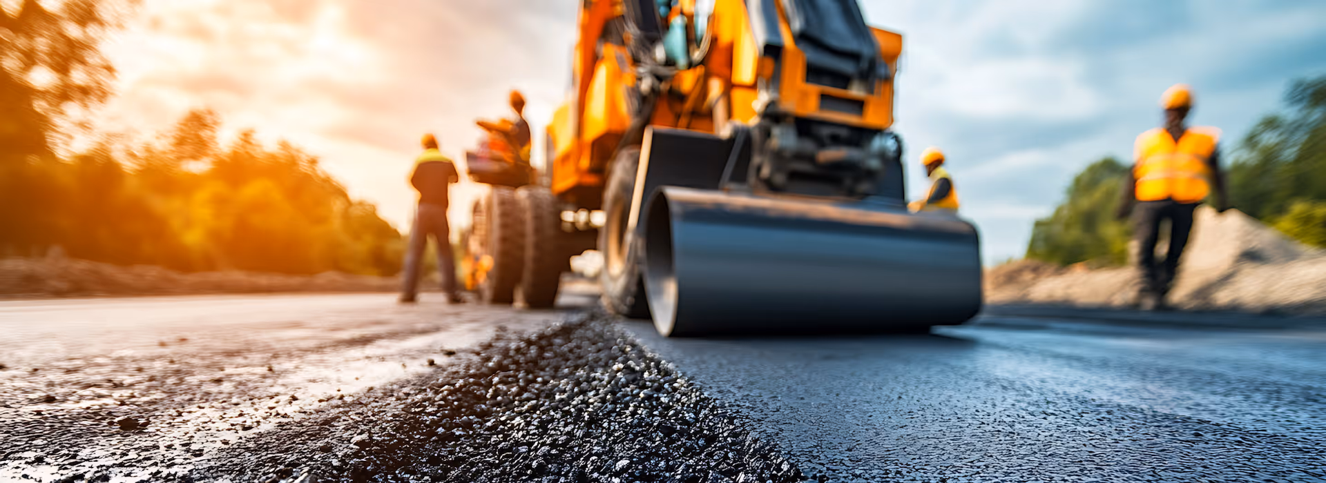 Road roller paving asphalt on a road with workers in orange safety vests and helmets.