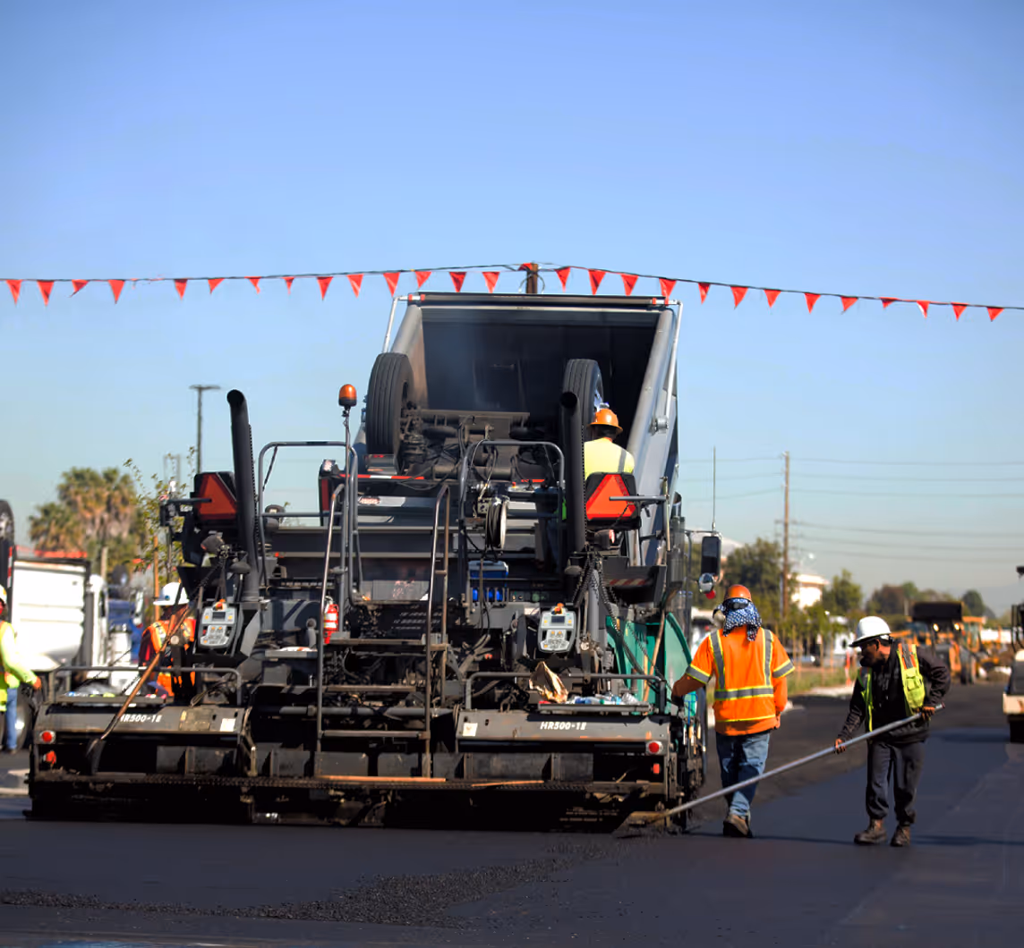 Workers wearing safety vests and helmets operating a large paving machine to lay asphalt on a road under clear blue sky.