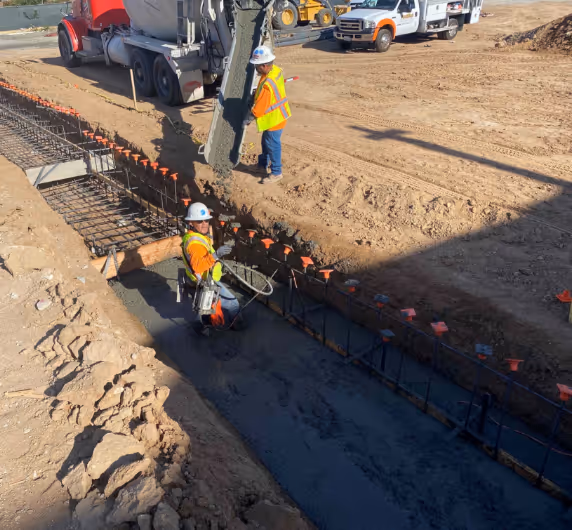 Two construction workers in safety gear pouring concrete into a trench at a construction site with trucks in the background.