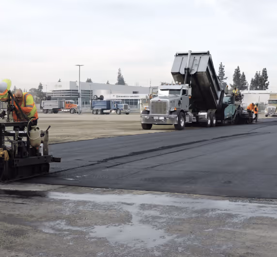 Workers in safety vests paving a road with asphalt using heavy machinery including a dumping truck and a paving machine.