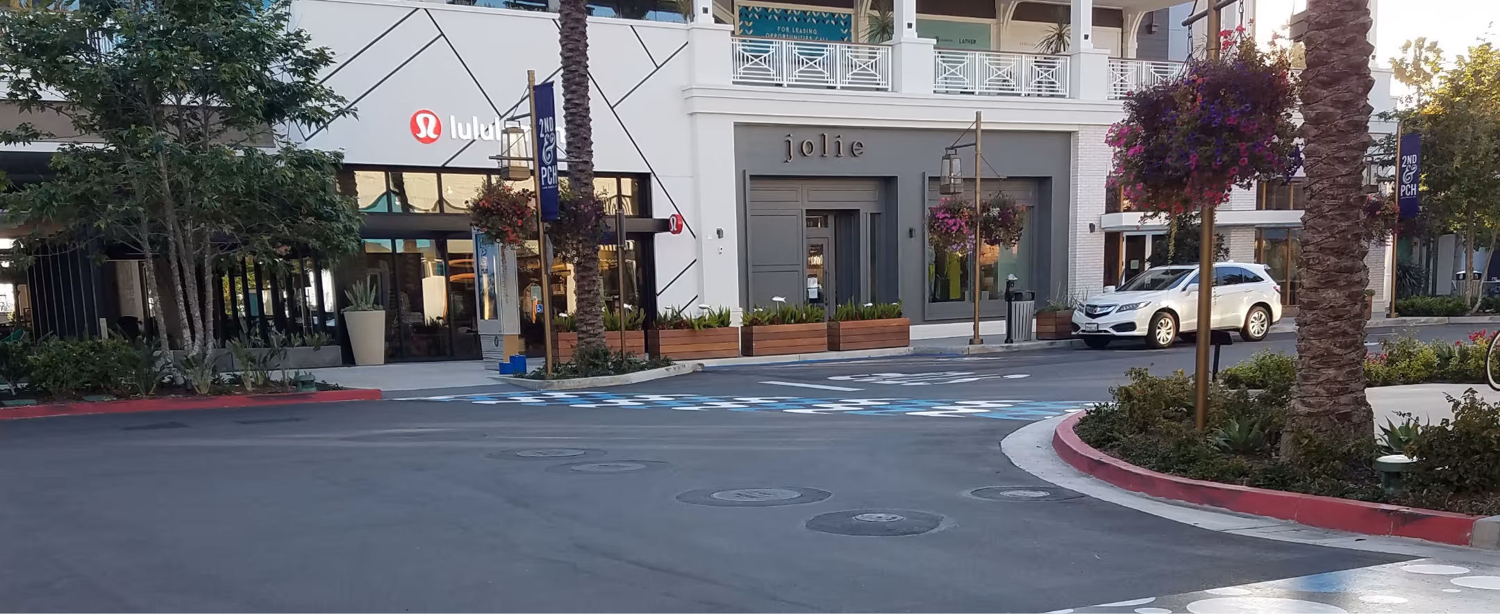 Outdoor shopping area with Lululemon and Jolie storefronts, palm trees, hanging flower baskets, and a white SUV parked nearby.