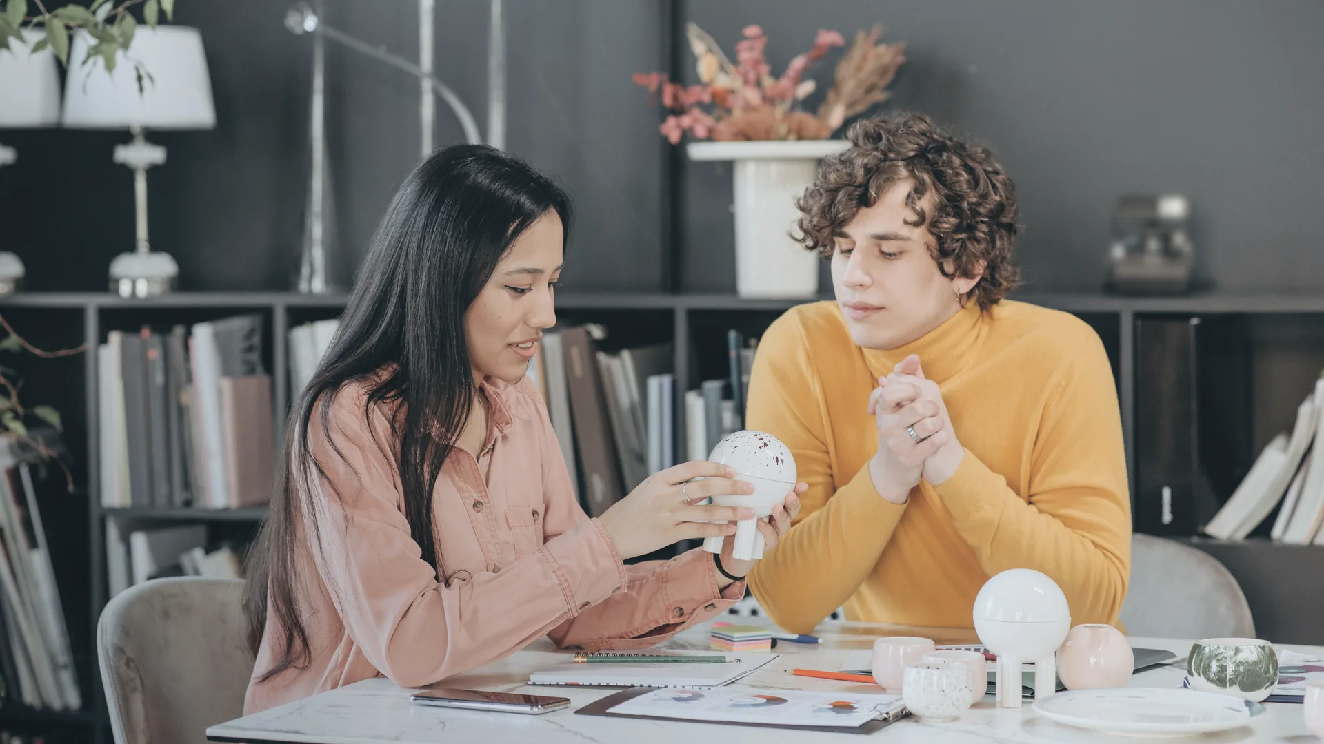 A brand founder presenting a product sample to a retail buyer across a table covered with samples and printed sheets.