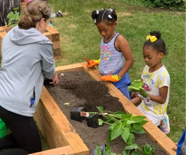 Two children with gardening gloves and an adult planting in a raised garden bed outdoors.