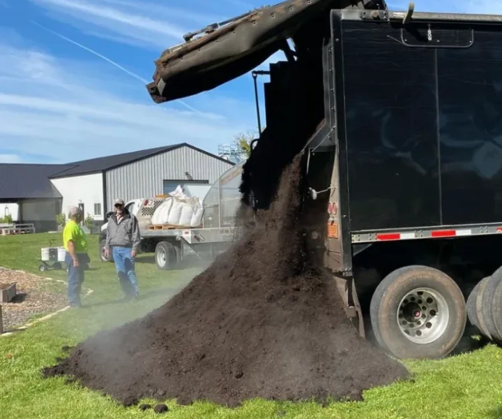 Black truck dumping a large pile of dark soil onto grass with two men standing nearby outdoors.