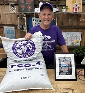 Smiling man in a purple cap and shirt holding a large bag of PCO1 Cow Organics and an award on the table.