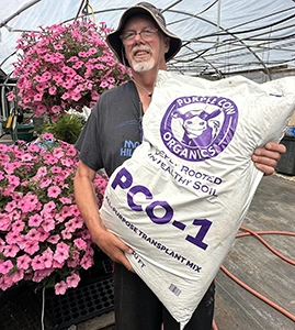 Smiling man in hat holding a large bag of PCO-1 organic soil mix in a greenhouse with pink flowers.