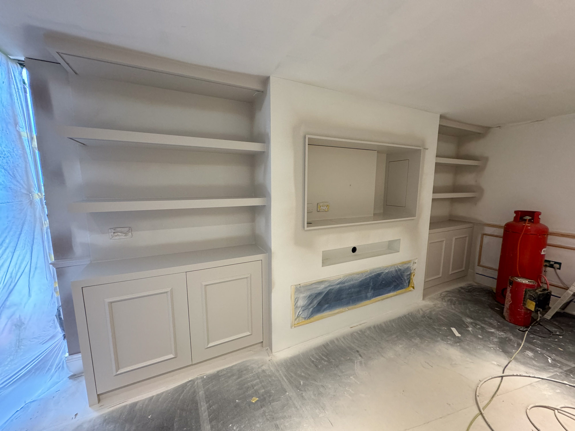 A white room under renovation with built-in shelves and cabinets, a covered fireplace, and construction equipment on the floor.