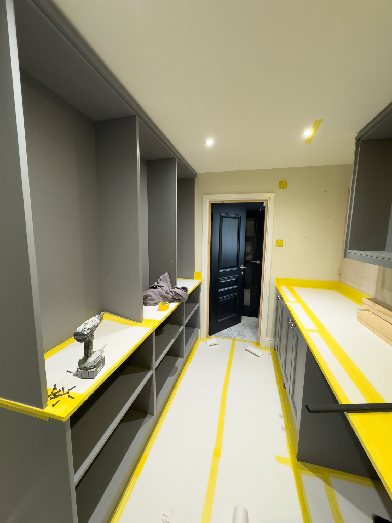 Interior of a narrow room with gray cabinetry and white countertops covered in protective paper with yellow tape, tools and screws on the left counter, and a black door open at the far end.