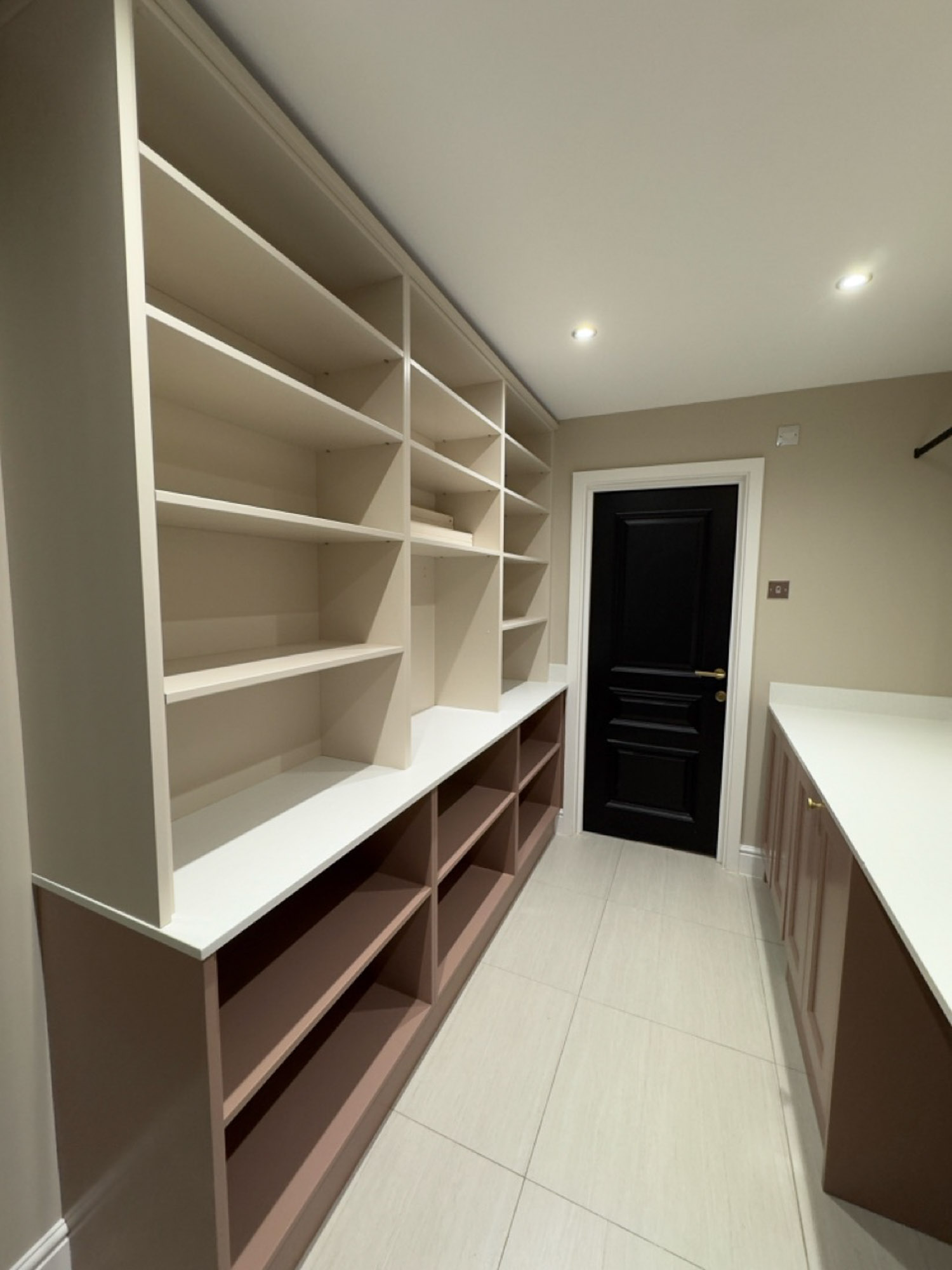 Modern pantry or storage room with built-in beige and brown shelves and cabinets, white countertops, tiled floor, and a black door.