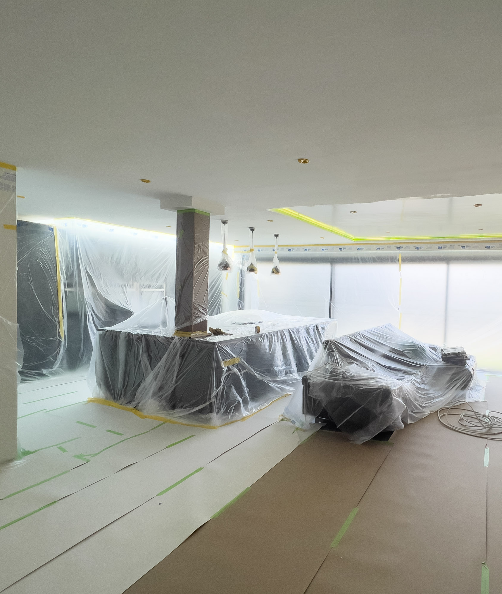 Interior living space under renovation with furniture and fixtures covered in protective plastic sheeting and floor covered with paper and tape.