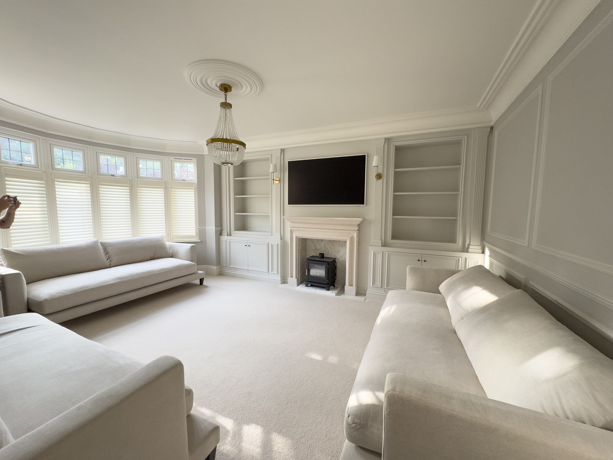 Bright living room with two cream sofas, large windows with shutters, a chandelier, and a fireplace with a mounted TV above.