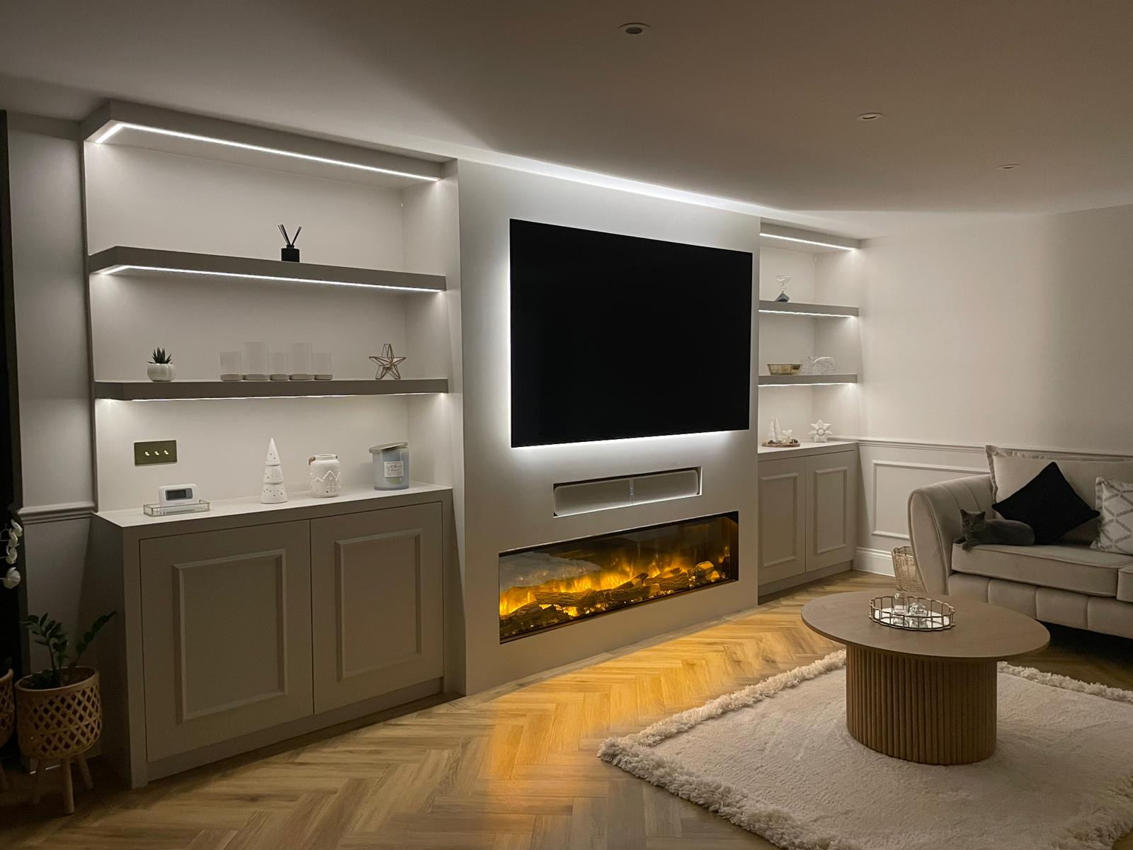 Modern living room with built-in illuminated shelves, wall-mounted TV above an electric fireplace, beige sofa with a cat, and round wooden coffee table on a white rug.