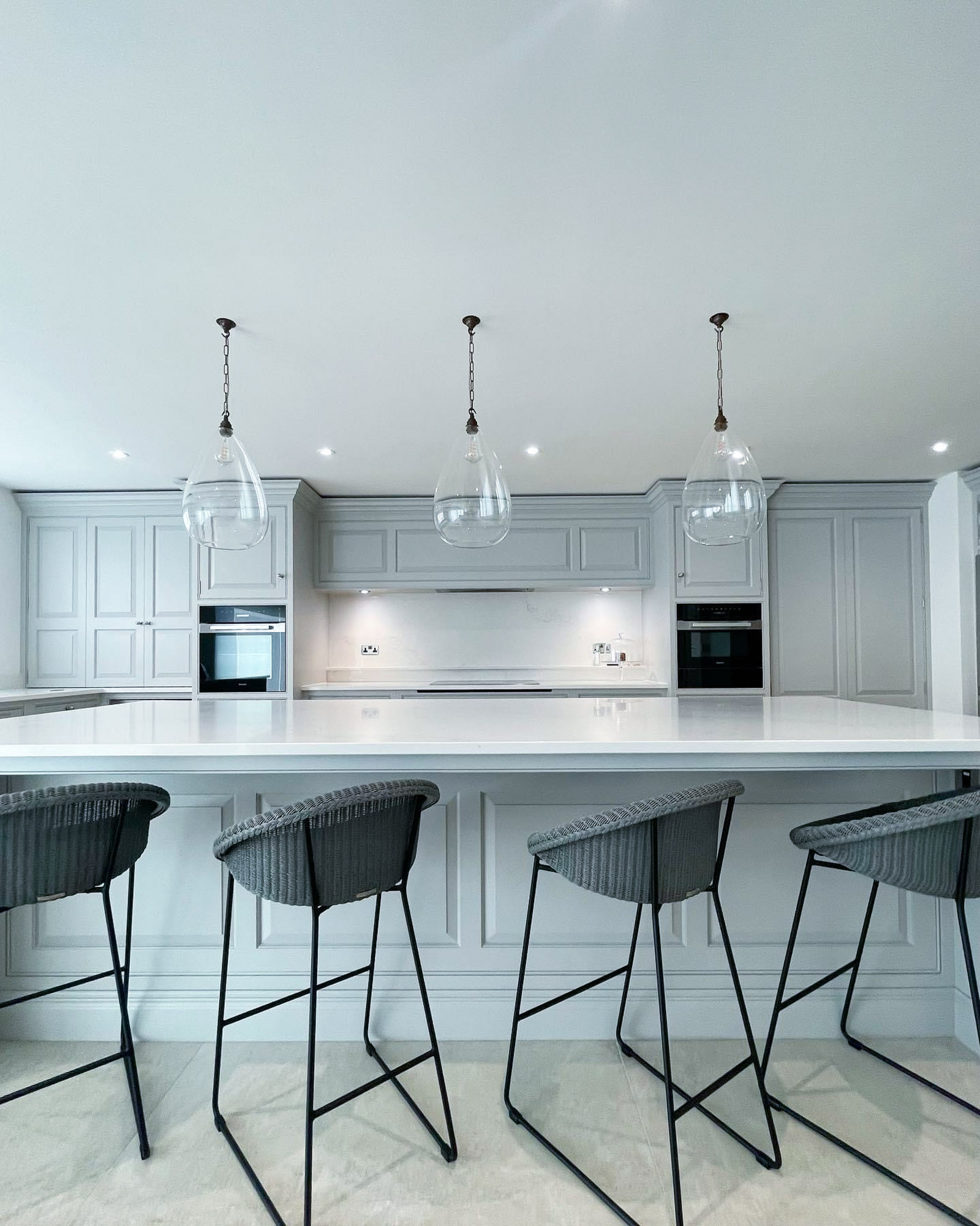 Modern white kitchen with large island, four gray bar stools, and three glass pendant lights overhead.