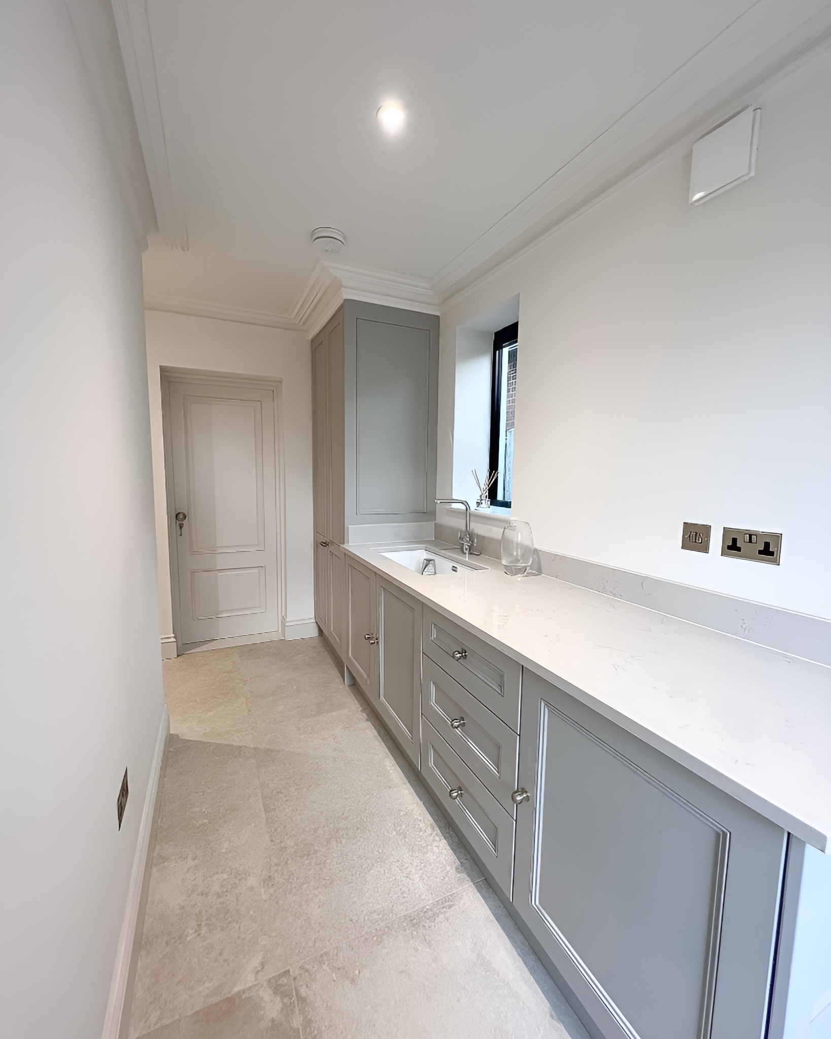 Narrow modern laundry room with gray cabinets, white countertop, built-in sink, small window, and beige tiled floor.