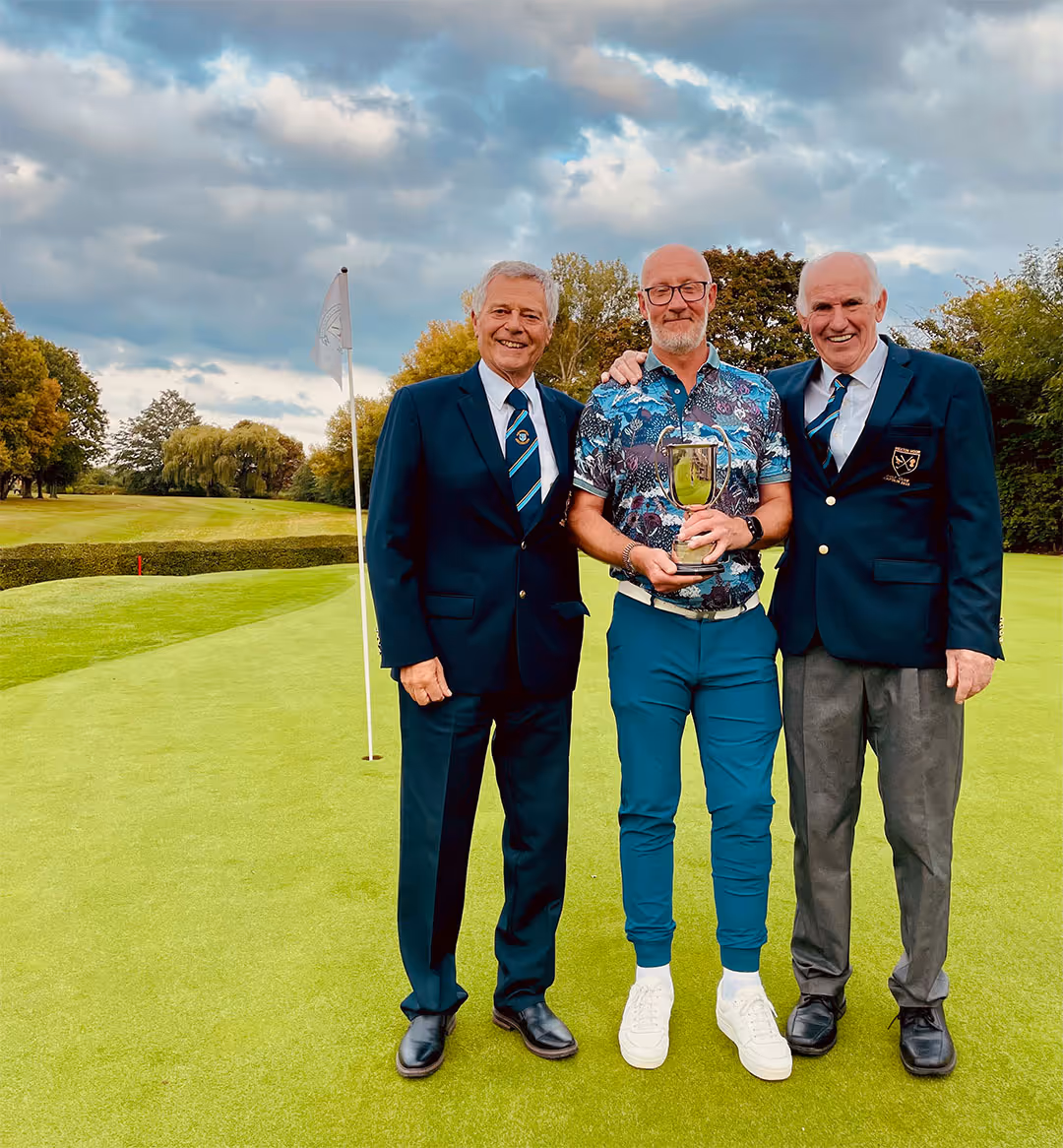 Three men standing on a golf green with a flag, the man in the center holding a trophy and smiling.