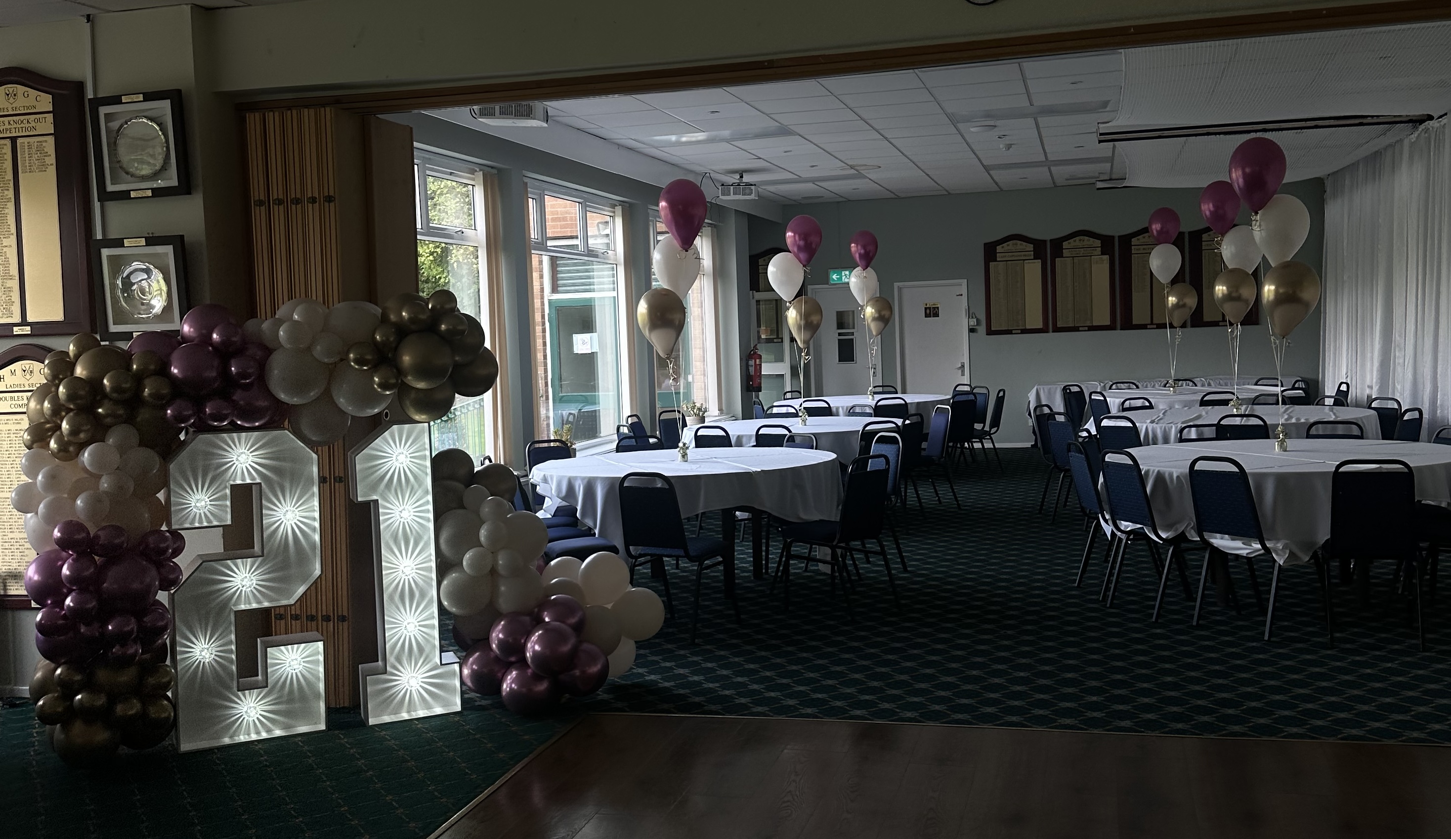Indoor event room set up with round tables covered in white tablecloths, blue chairs, clusters of white, gold, and purple balloons, and illuminated number balloons forming '21'.