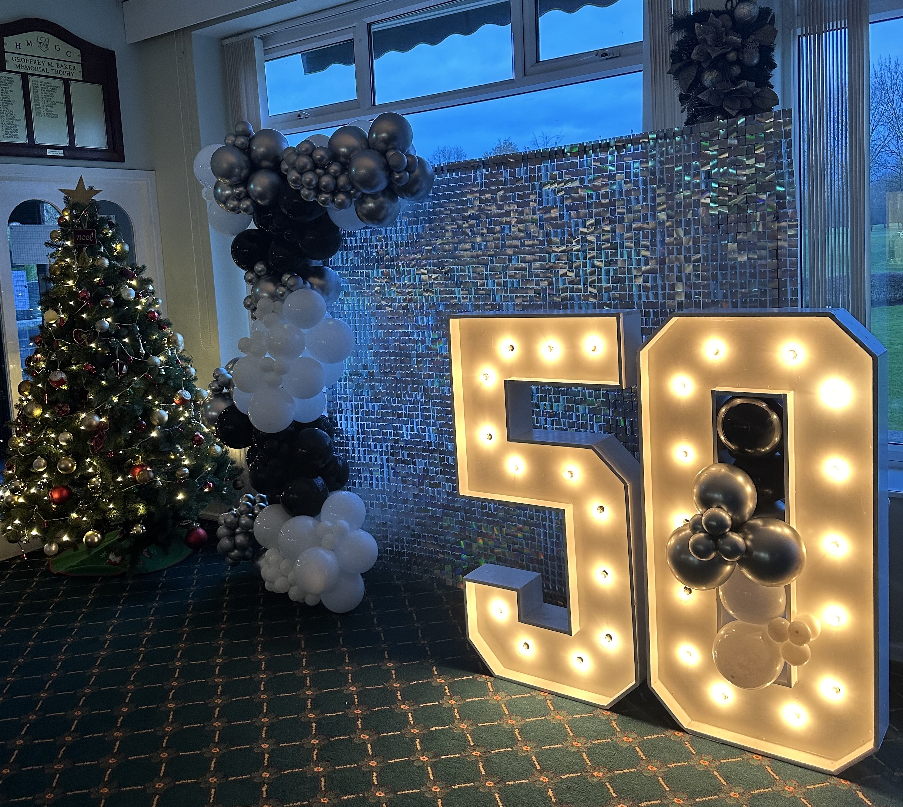 Indoor celebration setup with a decorated Christmas tree, black and white balloon arch, and illuminated number 50.
