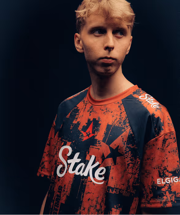 Young man with blonde hair wearing a black and red 'Stake' shirt against a dark background.