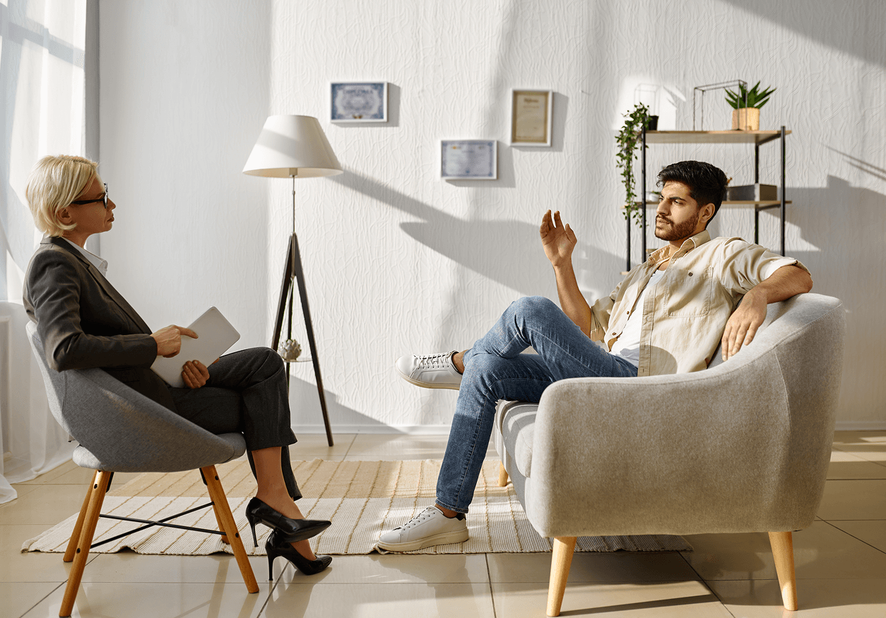 Man in casual clothes talking while seated on a sofa during a therapy session with a female therapist holding a tablet.