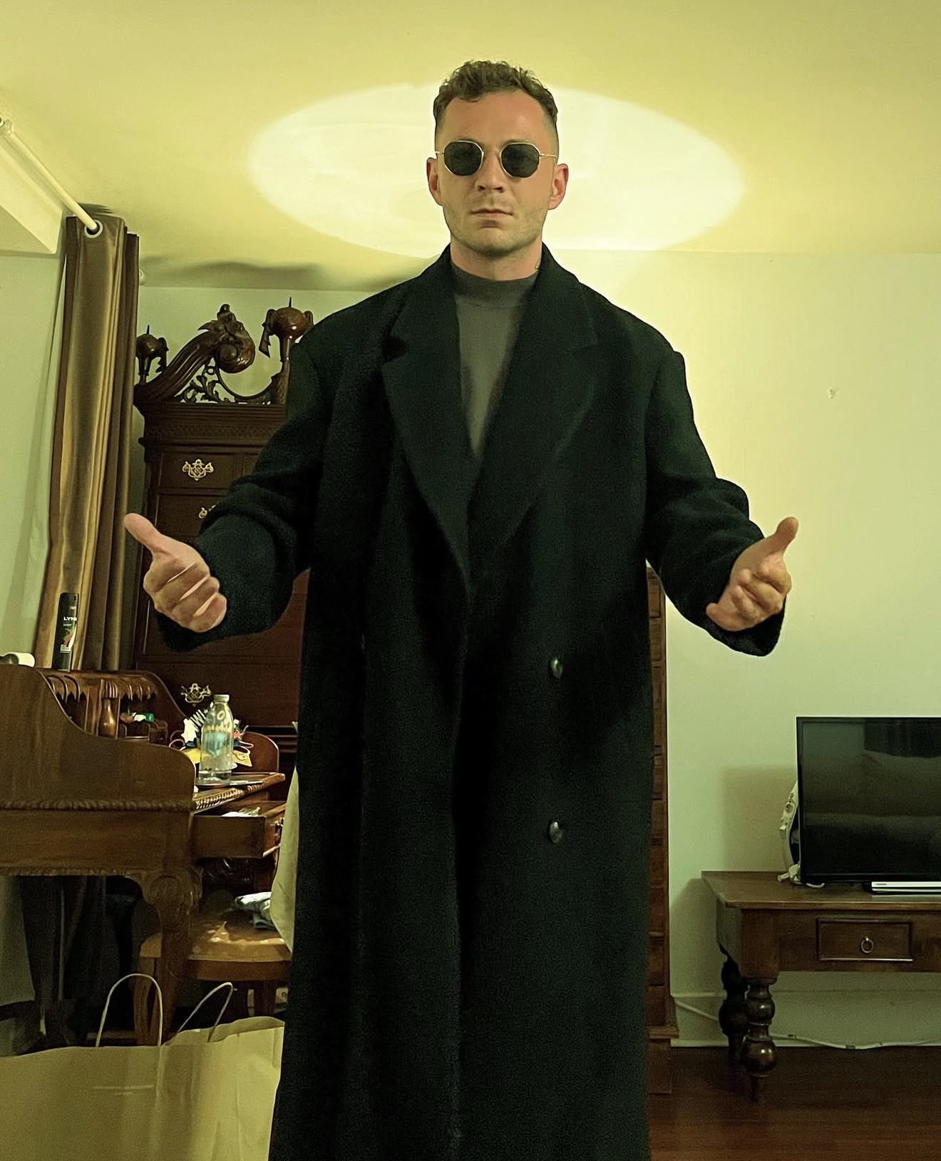 Man wearing a long black coat and round sunglasses standing with open arms in a living room.