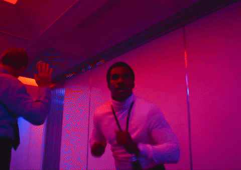 Man in white turtleneck jacket making quick movements in a pink and purple lit room with another person raising their hand on the left.