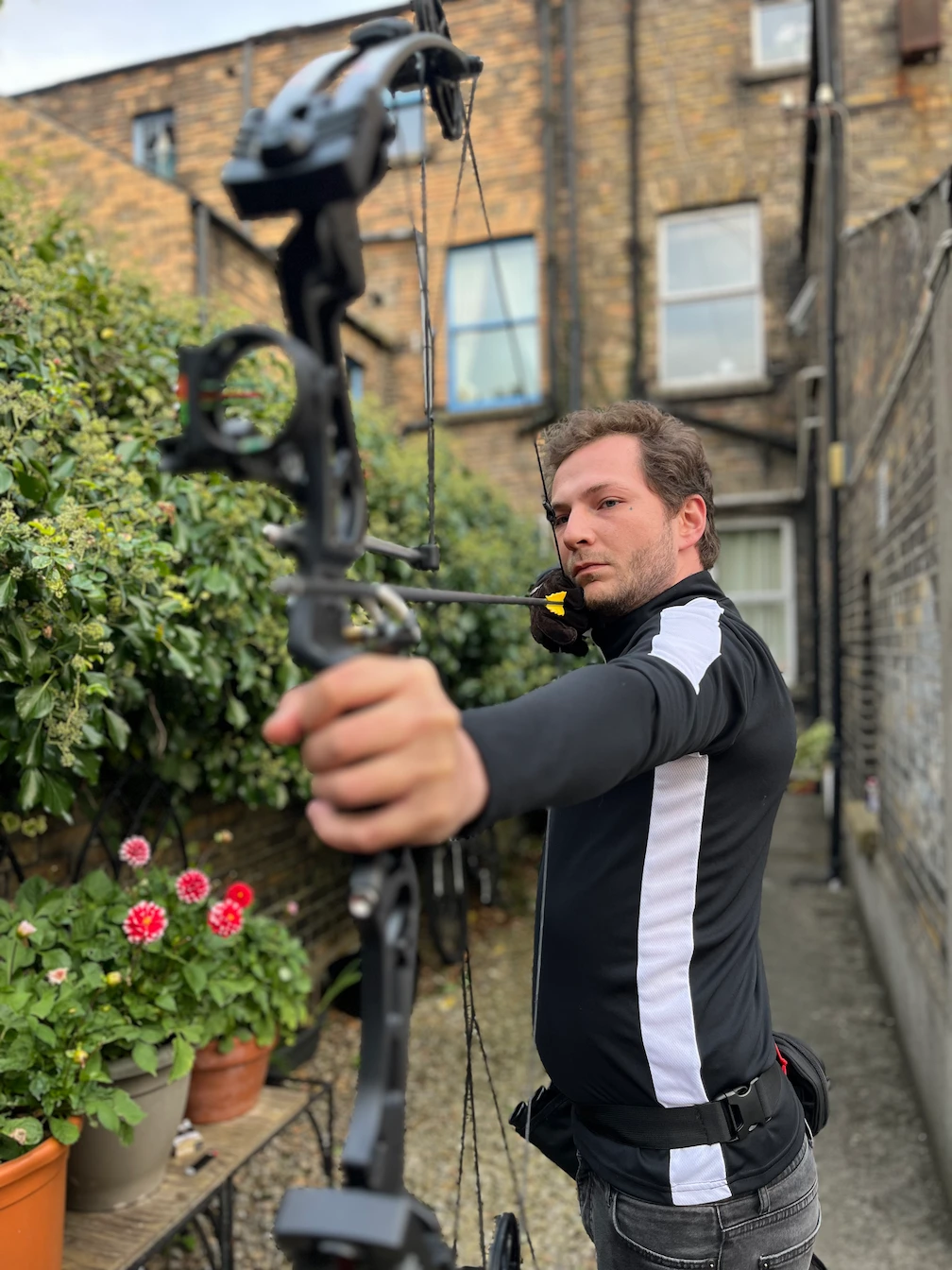 Man aiming a black compound bow with an arrow in an outdoor garden area.