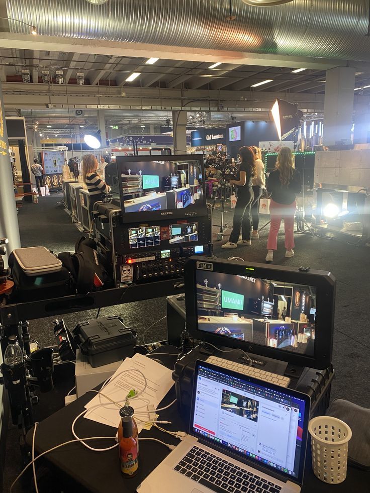 Control station with monitors, laptop, and camera equipment during a live event production inside a large indoor venue with people in the background.