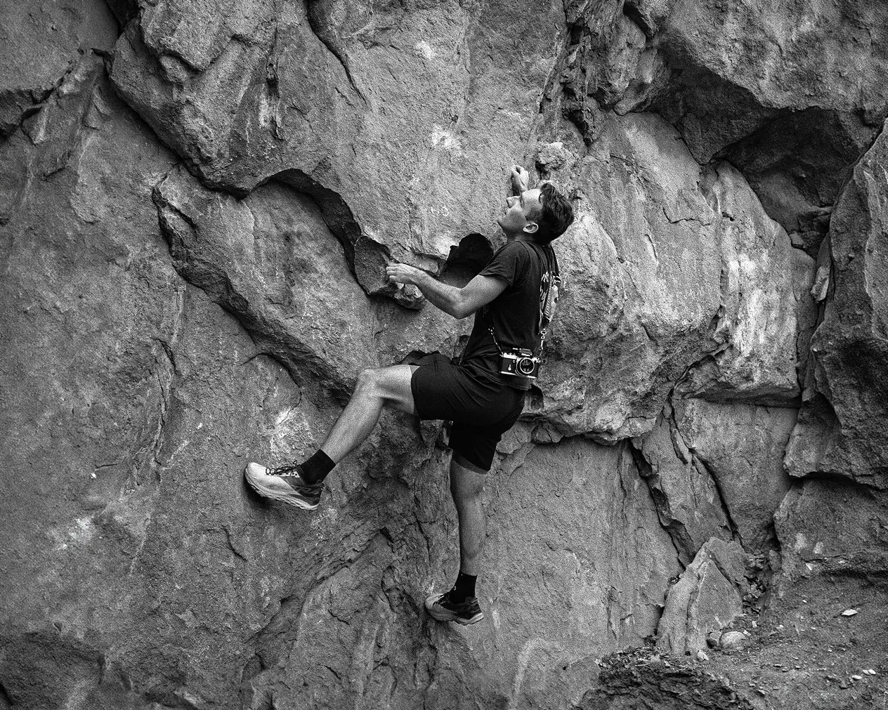 Black-and-white photo of Matt O’Donnell climbing a rock face, a camera slung across his chest.