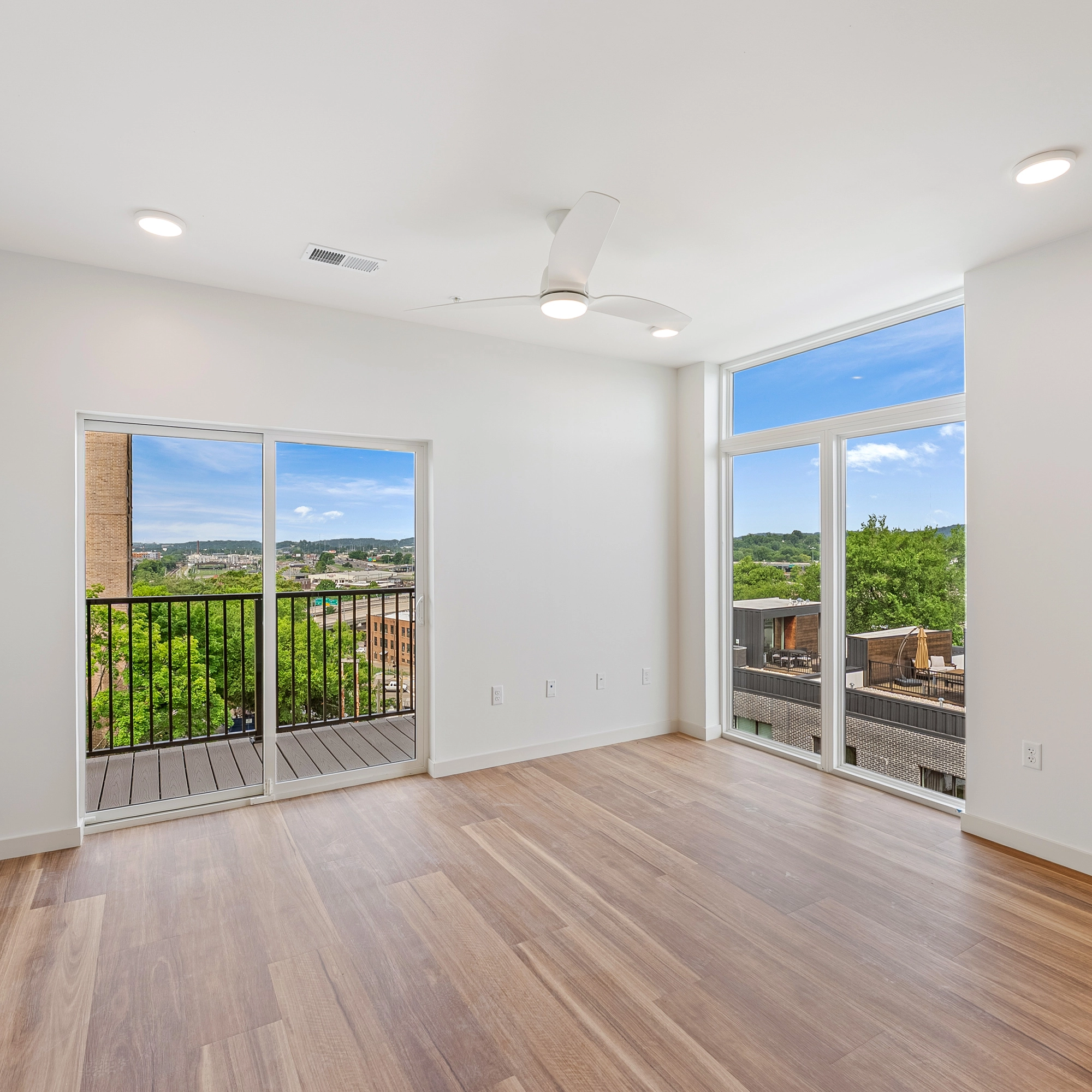Empty modern room with wood floor, ceiling fan, balcony door, and large windows showing cityscape.
