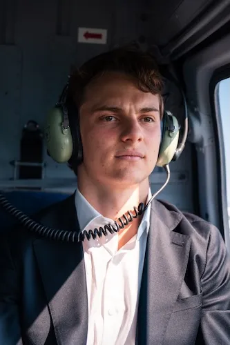 Oscar Piastri in suit wearing a headset inside a vehicle, looking out the window.