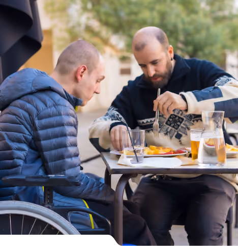 Two men sharing a meal at an outdoor table, one seated in a wheelchair