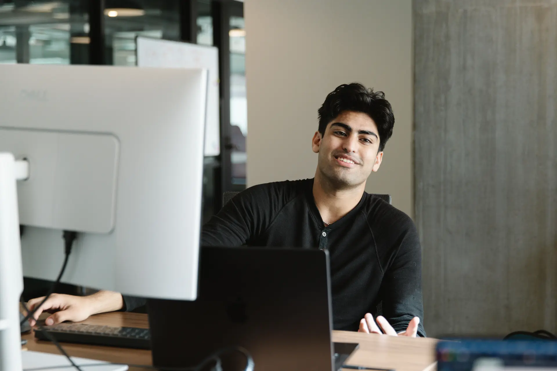 Smiling young man in black shirt sitting at a desk with multiple computer screens in an office.