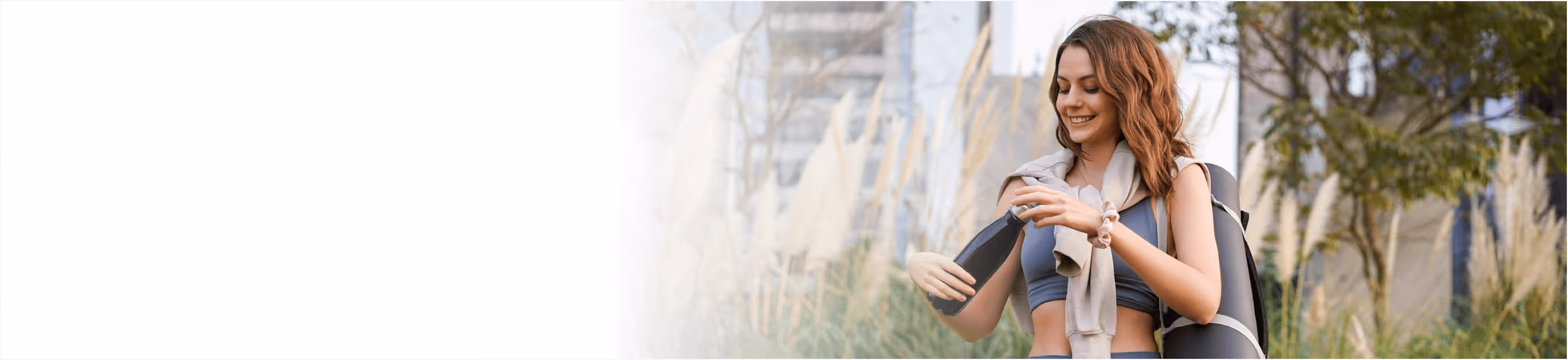 Smiling woman in workout clothes holding a water bottle and carrying a yoga mat outdoors.