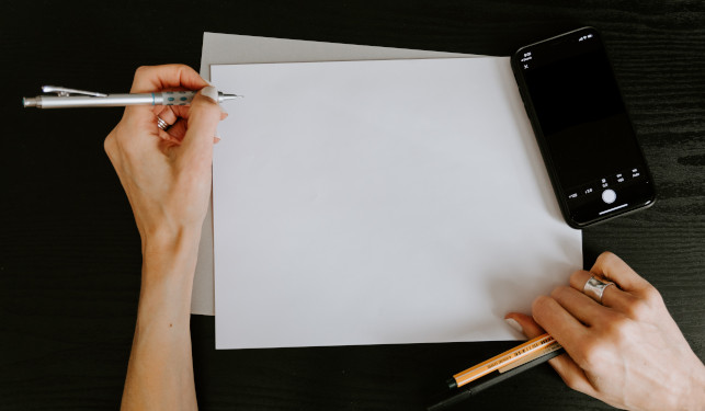 White paper against black background surrounded by a mobile phone and two hands holding pens