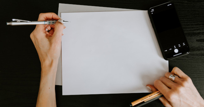 White paper against black background surrounded by a mobile phone and two hands holding pens
