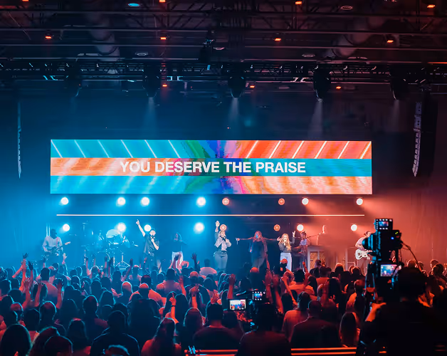 Concert stage with musicians and singers performing in front of a large crowd, a screen above displays the text 'YOU DESERVE THE PRAISE'.