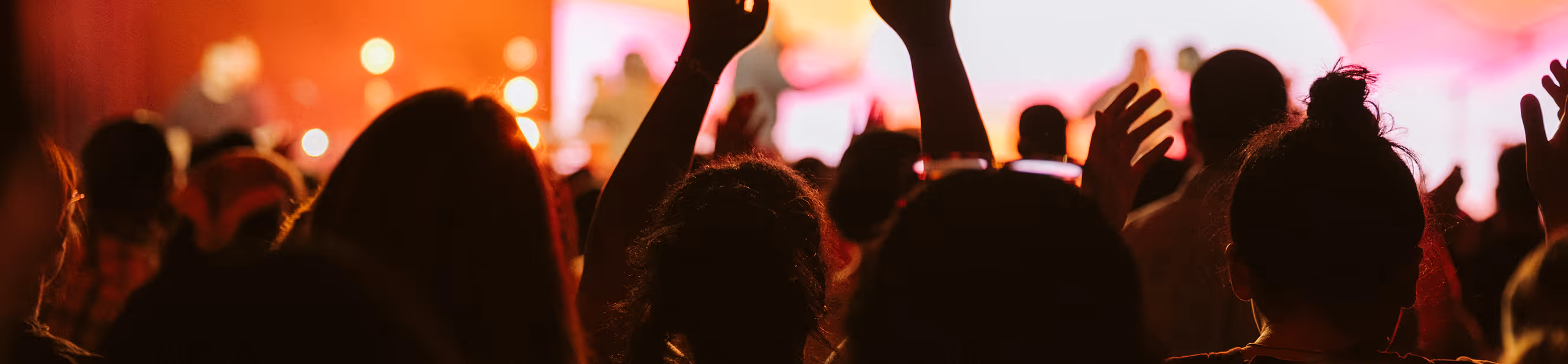 Crowd of people with raised hands enjoying a concert with warm, colorful stage lighting.