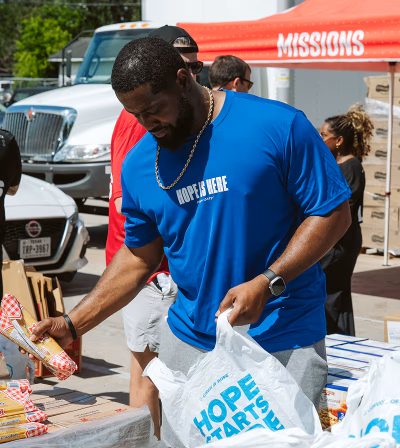 Man in blue 'Hope is Here' shirt placing packaged spaghetti into a plastic bag reading 'Hope Starts Here' at an outdoor event.