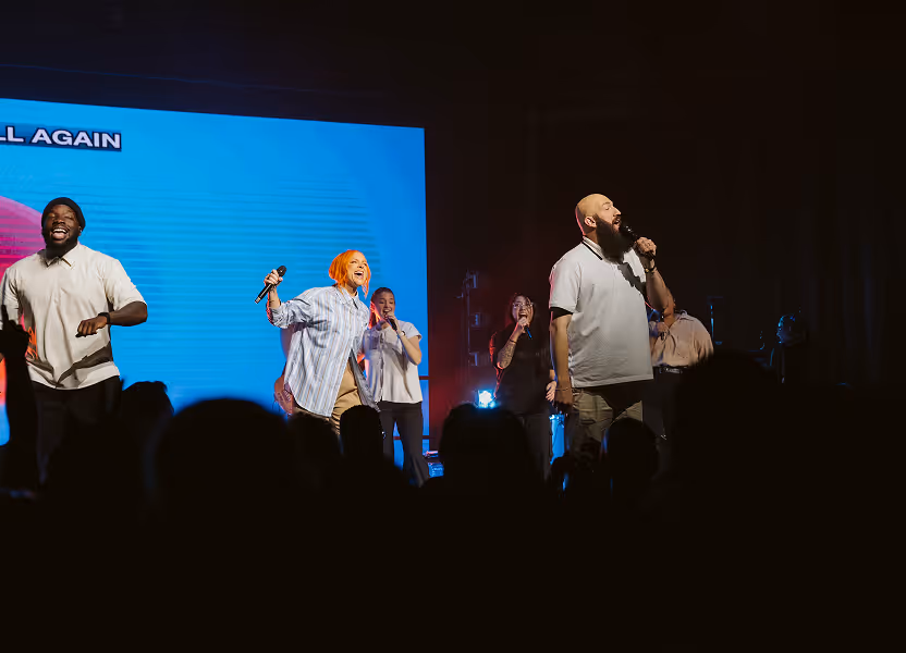 Group of five diverse singers performing on stage with microphones in front of an audience, illuminated by stage lights and a large blue screen backdrop.