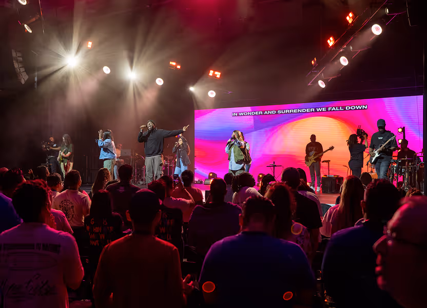 Crowded music concert with a band performing on a brightly lit stage and colorful LED screen displaying song lyrics.