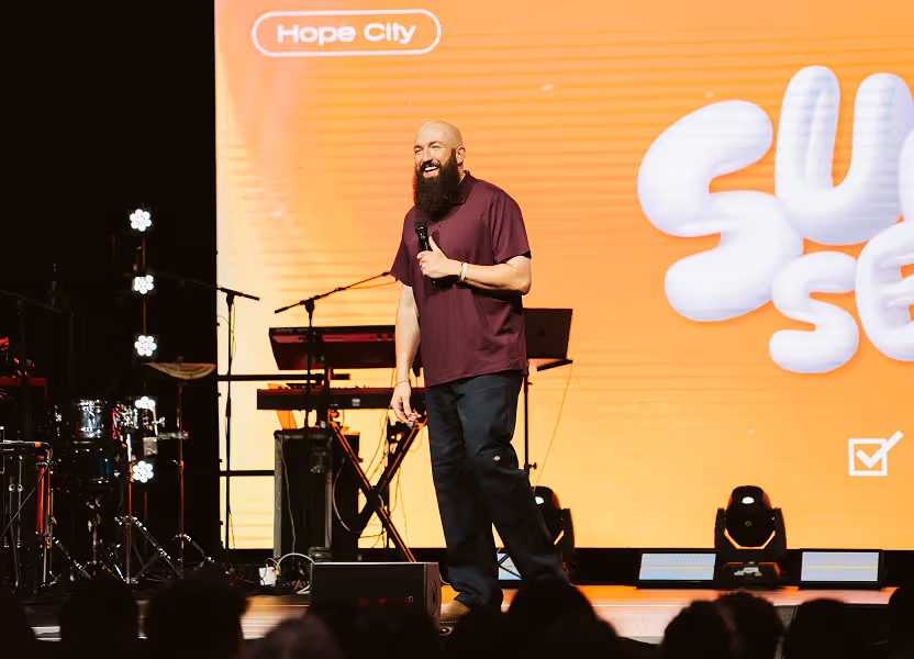 Bearded man smiling and speaking on stage with a microphone in front of an orange backdrop displaying the words 'Hope City' and partial text 'su se'.