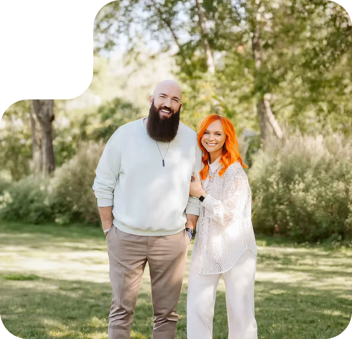 Smiling couple outdoors, man with a long beard wearing a white sweatshirt, woman with bright orange hair in a white lace top.