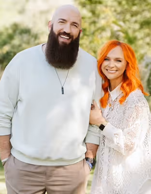 Smiling man with a beard wearing a white sweater and a woman with long red hair in a white lace dress posing outdoors with greenery background.