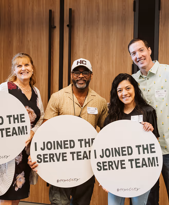 Four smiling adults holding round signs that say 'I JOINED THE SERVE TEAM! #hopecity' in front of wooden double doors.