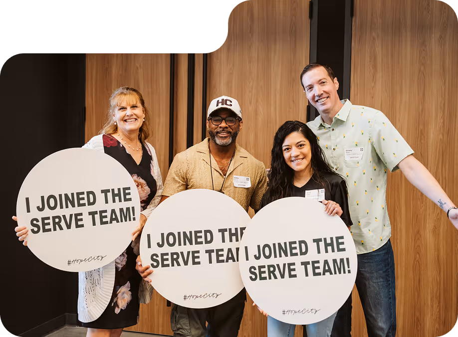 Four smiling adults holding round signs that say 'I JOINED THE SERVE TEAM! #hopecity' in front of wooden double doors.