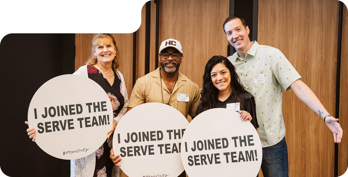 Four smiling adults holding round signs that say 'I JOINED THE SERVE TEAM! #hopecity' in front of wooden double doors.