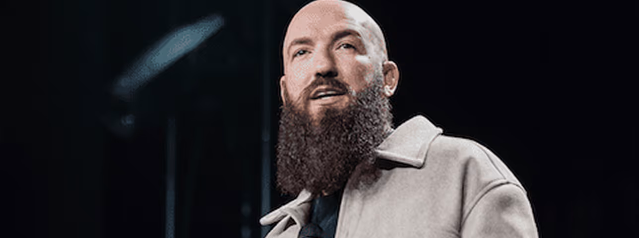 Bearded bald man wearing a beige coat looking slightly upwards against a dark background.