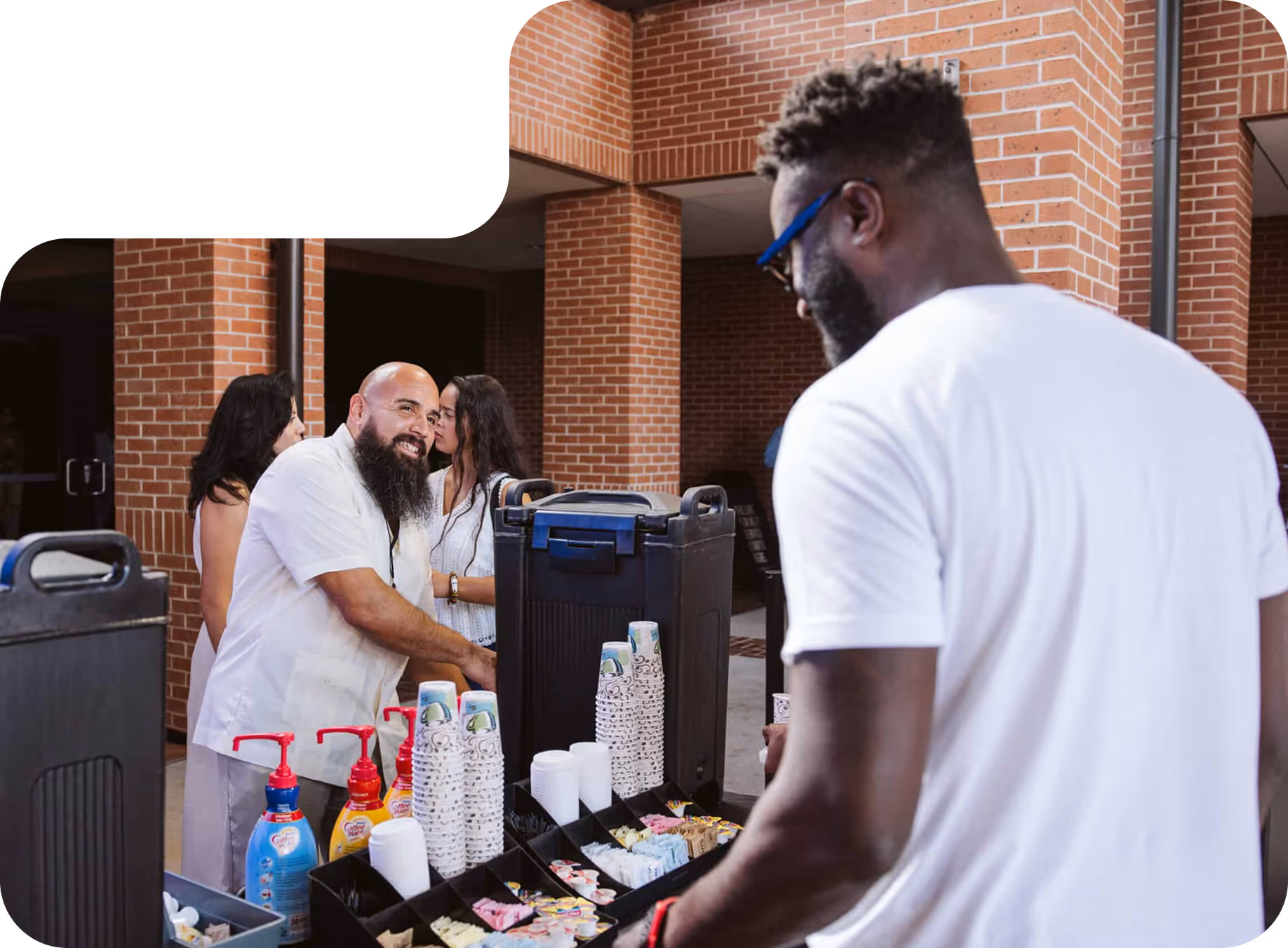 Two men at a coffee station with cups, sugar packets, and creamers, inside a brick building with other people in the background.